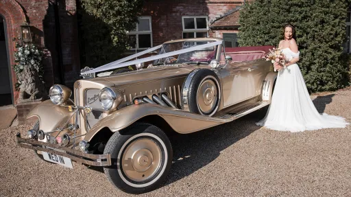 Bride in white wedding dress standing beside gold Beauford Convertible with chrome detailing and side-mounted spare wheel.