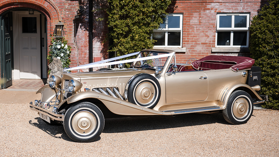 Side view of gold Beauford Convertible with burgundy interior parked outside a country wedding venue in Wiltshire.