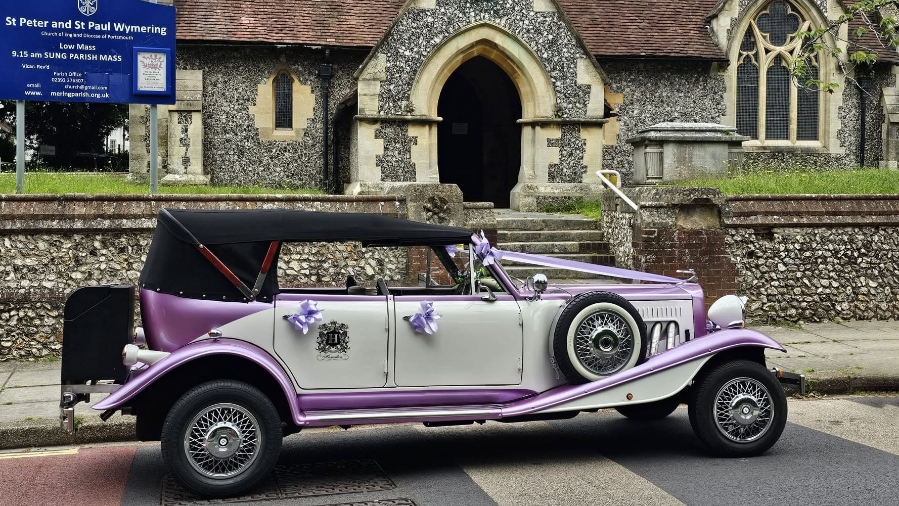 Right side view of the lilac and white four-door Beauford Convertible parked outside a church