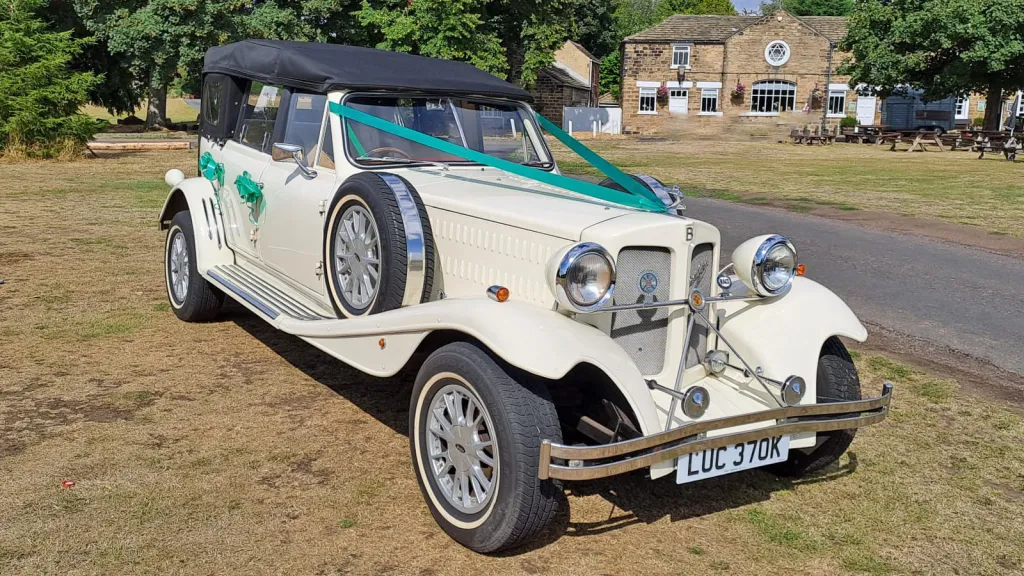Front three quarter view of Old English White four door Beauford Convertible wedding car with black soft top roof, dressed with green ribbons and bows and side mounted spare wheel