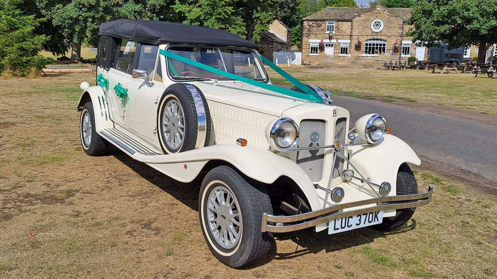 Front three quarter view of Old English White four door Beauford Convertible wedding car with black soft top roof, dressed with green ribbons and bows and side mounted spare wheel