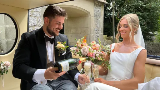 Groom pooring a glass of Champagne in the rear of a convertible vintage style wedding car to enjoy their journey together with cheerful music