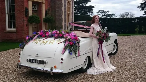 Classic Bentley with convertible roof open decorated with Pink flowers and Burgundy Ribbons. Bride wearing a white dress is standing beside the vehicle holding a matching bouquest of flowers in pink and burgundy.