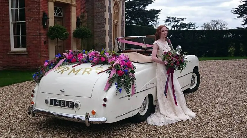 Classic Bentley with convertible roof open decorated with Pink flowers and Burgundy Ribbons. Bride wearing a white dress is standing beside the vehicle holding a matching bouquest of flowers in pink and burgundy.