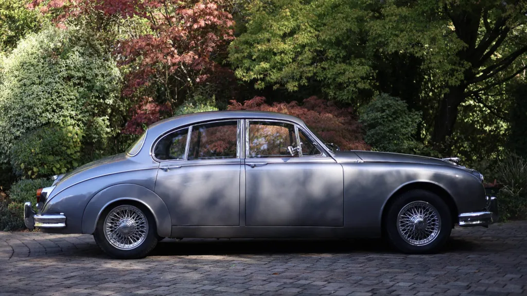 Full side profile of a silver classic Jaguar Mk2 parked beside trees
