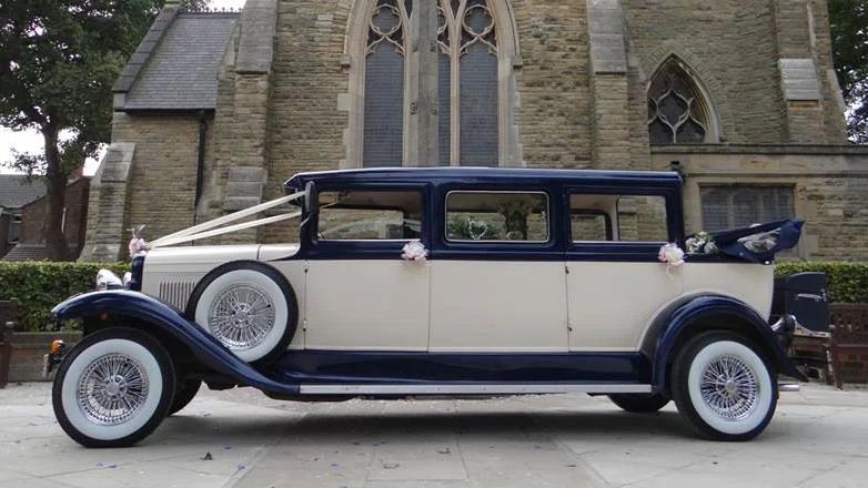 Side view of an ivory Bramwith Landaulette wedding car with a dark blue roof and wheel arches outside a church.