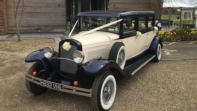 Front three-quarter view of an ivory and dark blue Bramwith Landaulette wedding car with whitewall tyres.