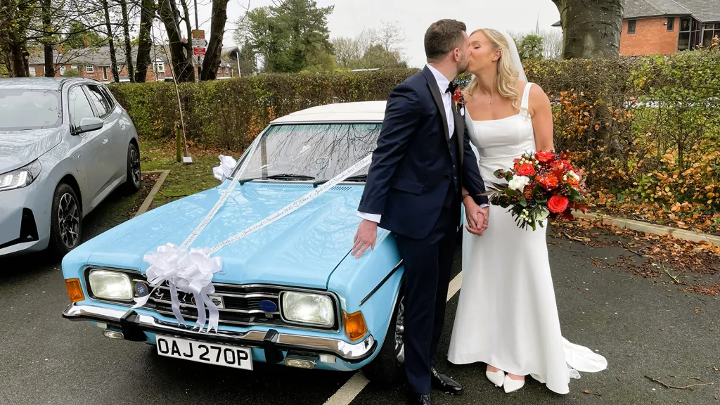 Bride and groom kissing beside a light blue Ford Cortina decorated with white wedding ribbons