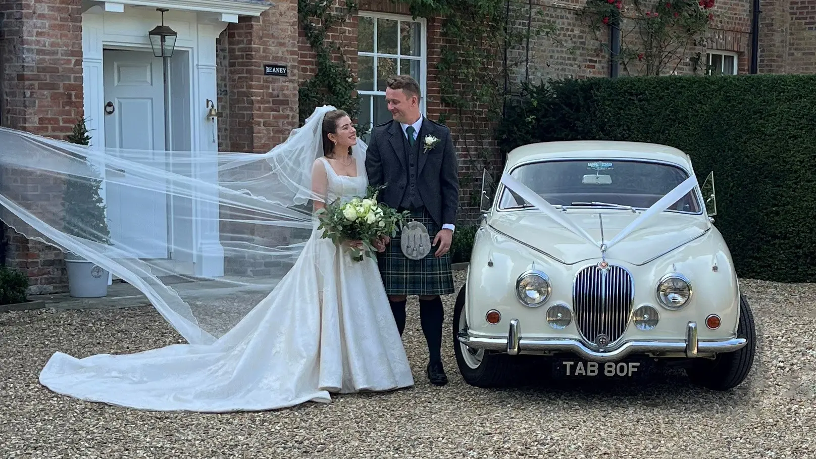 Bride and groom in a kilt beside an Old English White Jaguar MK2 wedding car outside a red-brick house, as the bride’s veil is held out for the photographer