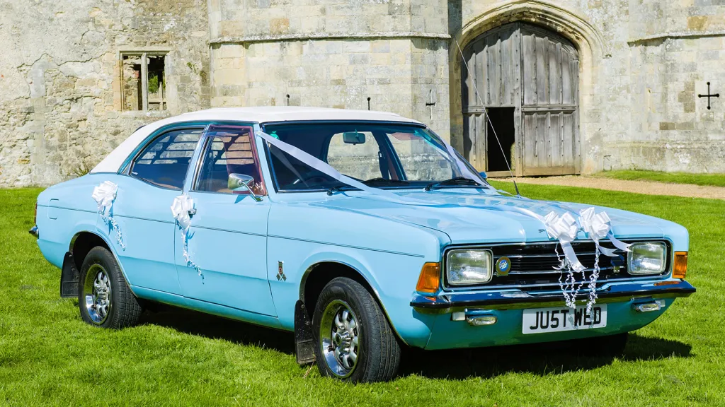 Light blue Ford Cortina Mk4 with white wedding ribbons parked outside a stone building