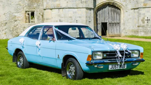 Light blue Ford Cortina Mk4 with white wedding ribbons parked outside a stone building