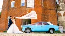 Bride and groom beside a light blue Ford Cortina with a white vinyl roof outside a brick building