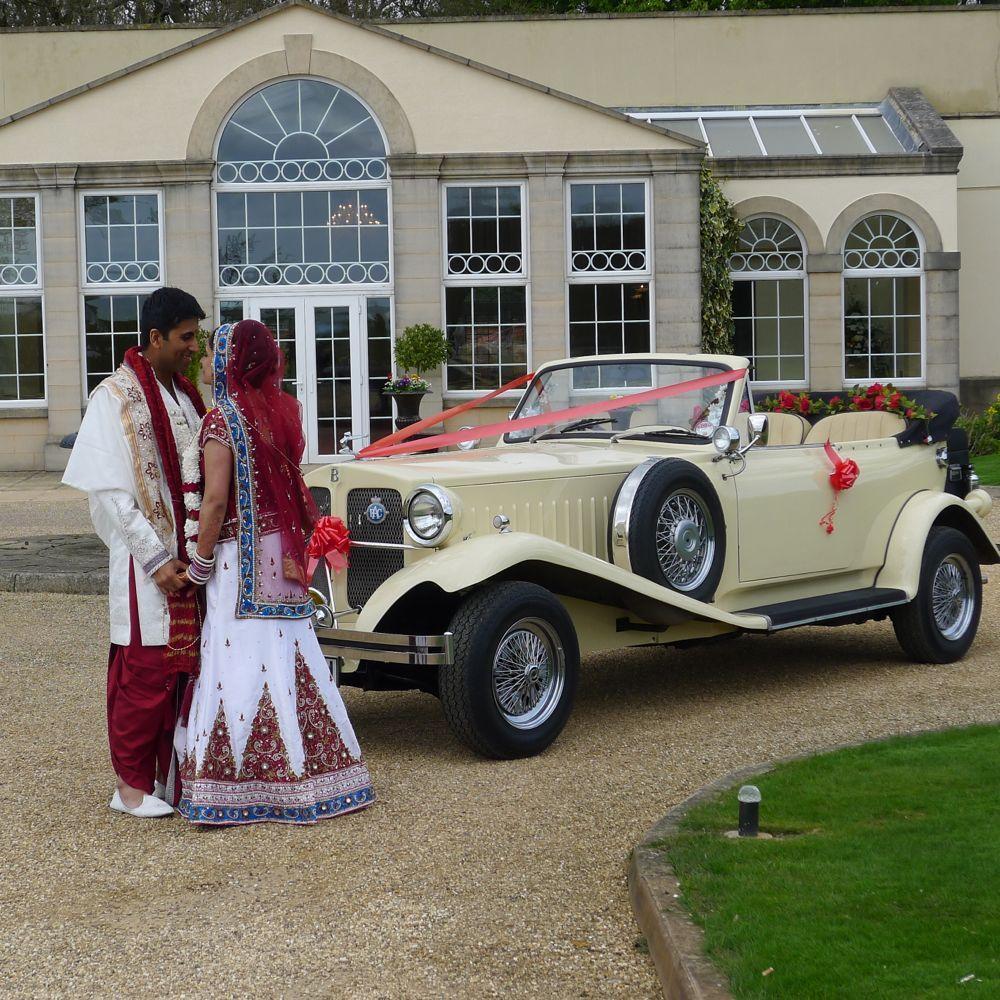 Ivory Vintage Beauford convertible with roof down and decorated with Red Ribbons. Asian couple are standing in front of the vehicle holding hands