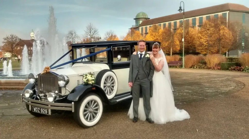 Vintage Imperial Convertible decorated with Dark Blue Ribbons and Gold bow at a West Midlands wedding venue. Bride and Groom are standing next to the vehicle smiling. 