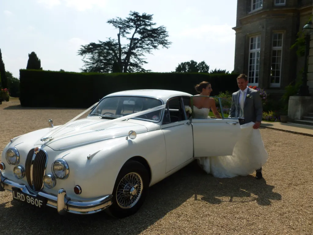 White classic Mk2 Jaguar decorated with white ribbons. Bride is exiting the vehicle while groom holds the door.