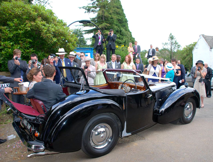 Black Triumph Convertible with Bride & Groom seating in the back seat and wedding guests standing in the background