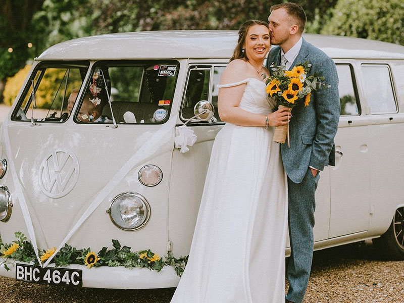 Classic Cream VW Campervan decorated with white ribbons and guarland of green and yellow flowers on its front bumper matching the Bridal bouquet. Couple standing by the vehicle with Groom kissing his smiling Bride