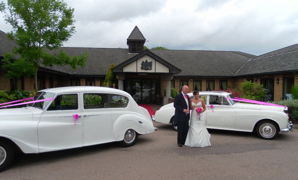 Two white classic cars with Bride and Groom standing in the middle. Both cars are decoratedwith matching Pink Ribbons accross the Bonnet and matching pink bows on the door handles. Pink colour matches the Bridal bouquet. 