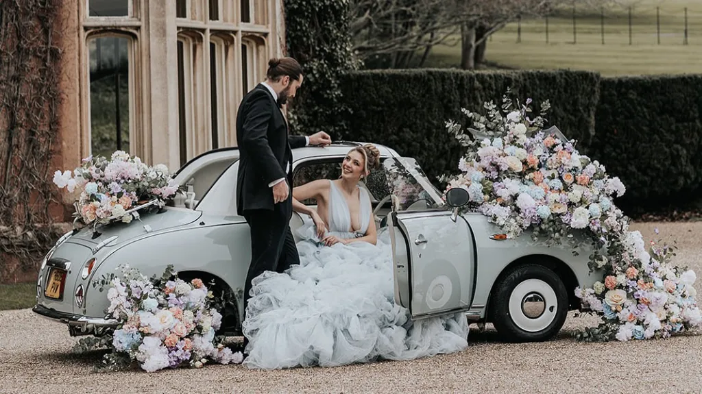 Classic Car decorated with a large display of white flowers on rear boot and front bonnet. Bride wearing a large white dress is seating in the vehicle smiling and looking at her Groom standing by the vehicle 