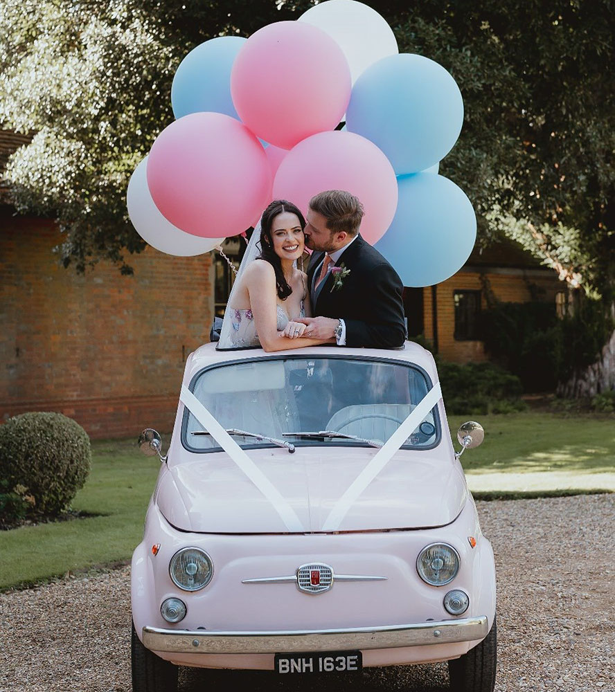Classic Pink convertible car with roof open and Bride and Groom standing inside the vehicle through the open roof. Car is decorated with white ribbons and Blue and Pink Balloons. 