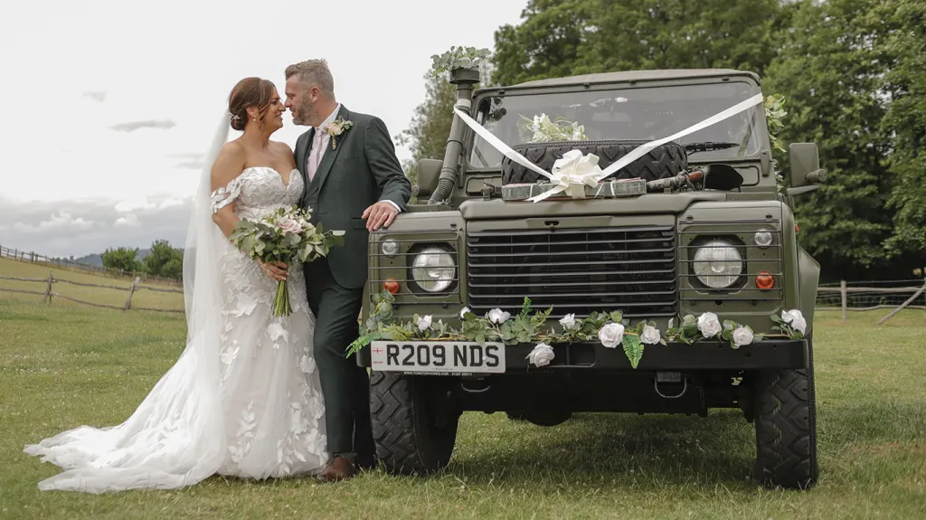 Khaki Green Landrover decorated with white ribbons and flowers on its front bumper. Bride and Groom are standing by the vehicle looking at each others. 