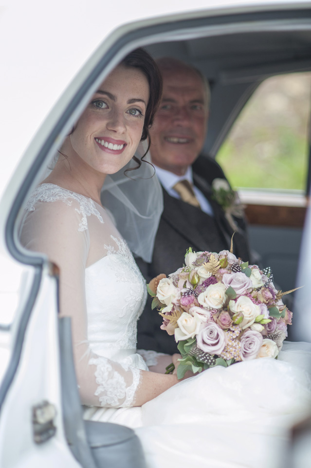 Bride and her father seating in the rear of a Classic Rolls-Royce. Bride is holding her bridal bouquet and both are looking at the photographer smiling. 