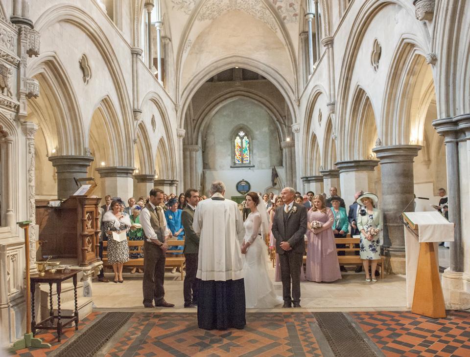 Inside Boxgrove church during the ceremony. Bride and Groom are standing in front of the priest and their wedding guests are seated in the background
