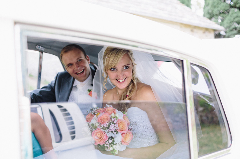 Bride and Groom inside Classic Rolls-Royce. Both smiling looking a their wedding guests through the window