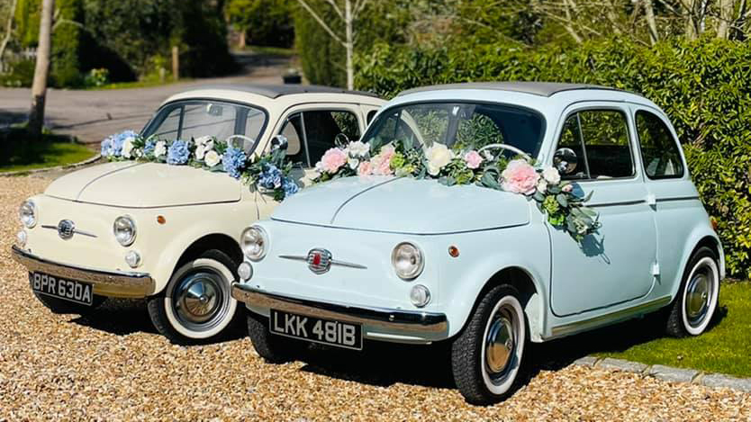 Two classic Fiat in Cream and pale blue decorated with flowers across their bonnets. 