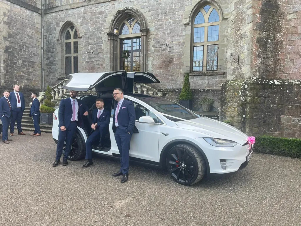White tesla X with Gull wings open parked in front of the wedding venue. Groom and his bestmen having photos taken