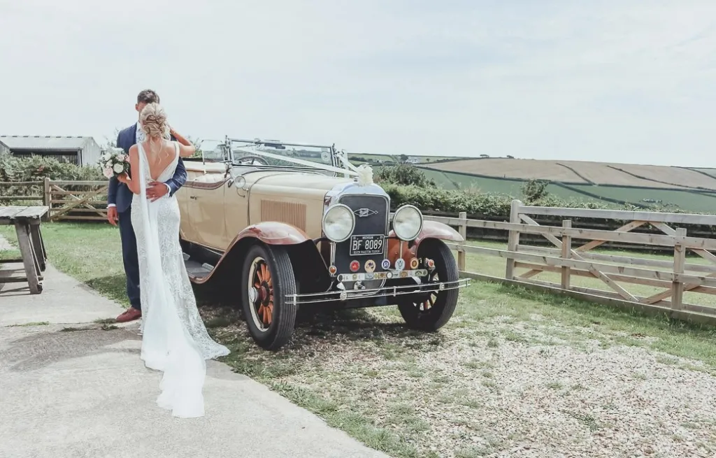 Groom holding Bride at the Barn Wedding Venue with Vintage Buick decorated with white ribbons in the background. 
