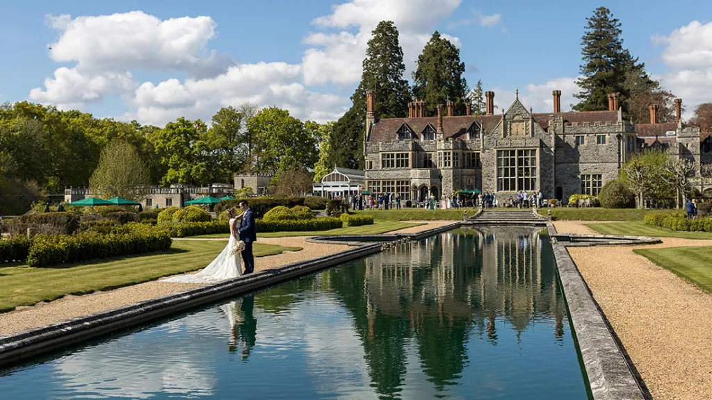 rear garden at Rhinefield House. Bride and Groom standing by the pond holding each others while wedding guests can be seen in the far background