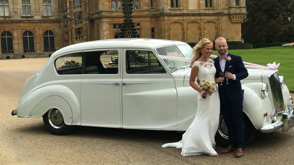 White classic Austin princess limousine with pale pink ribbons. Bride and groom are standing in front of the vehicle holding a glass of Champagne