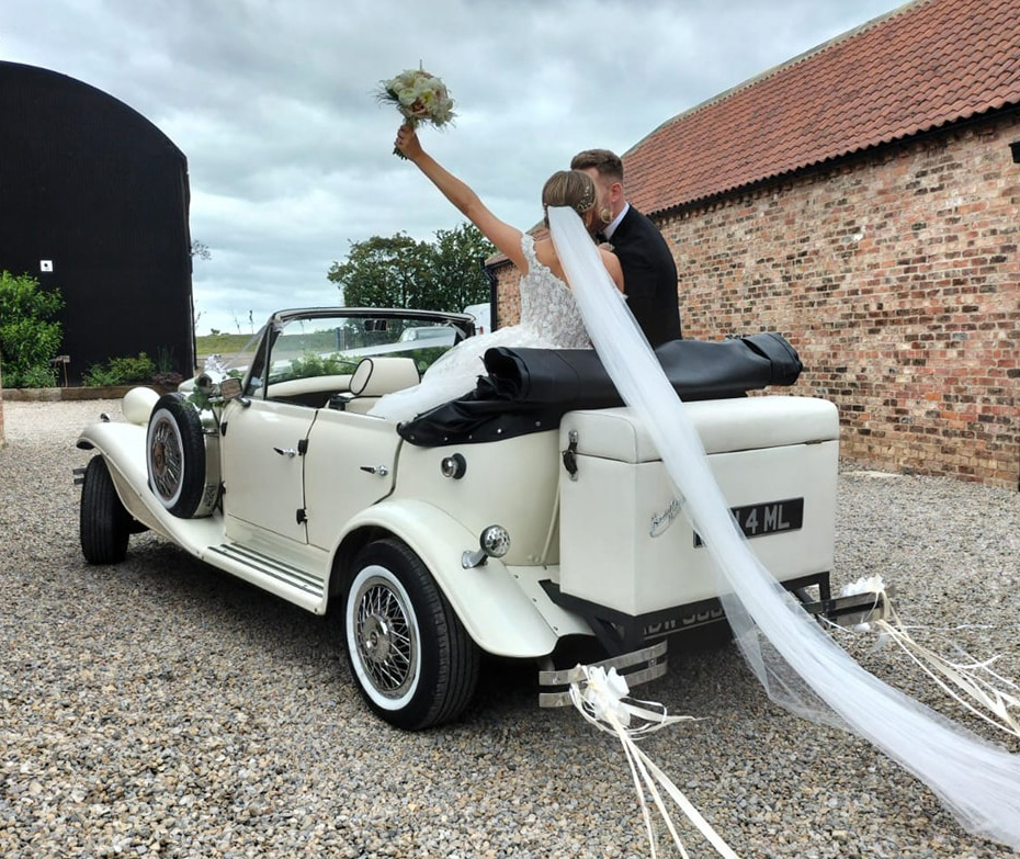 Ivory convertible Beauford with roof down. Bride and Groom are seated in the vehicle kissing each others. Bride is holding a flower of bouquet high up above her head. 