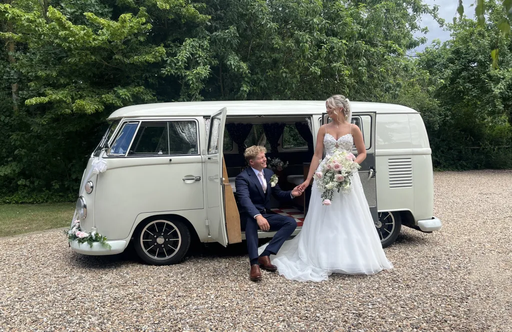 Classic Ivory Retro Campervan decorated with green foliage on front bumper. Groom is seated on the rear entrance and Bride is standing next to him