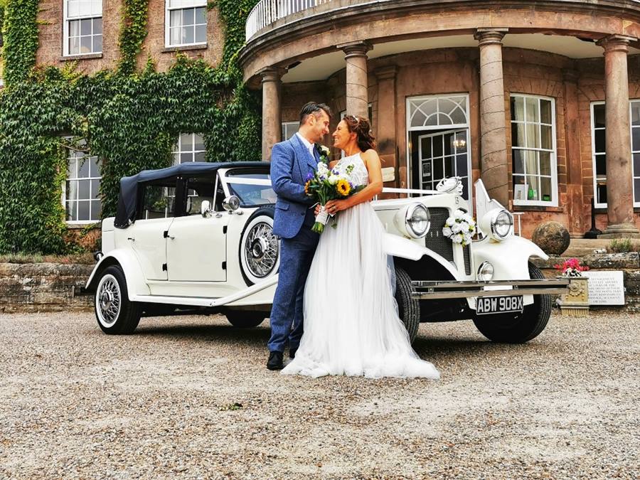 1930s vintage style white Beauford with black soft-top close. Bride and Groom are standing in front of the vehicle smiling at each others. 