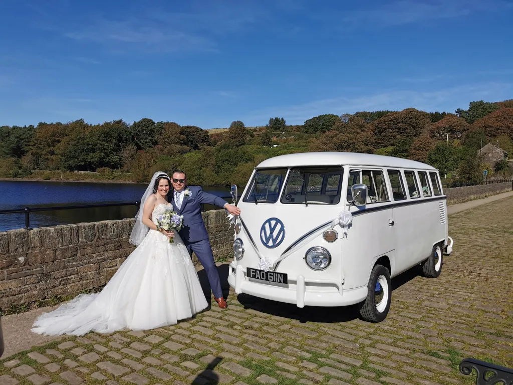 Classic splitscreen campervan with Bride and Groom standing by the vehicle for photos. Blue lac in the background.