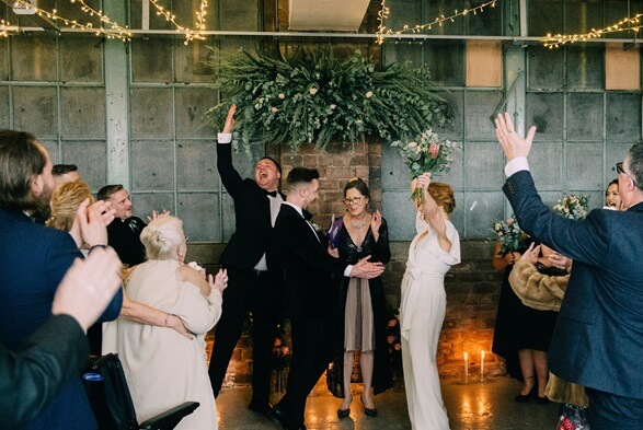 couple cheer with friends and family clapping in the foreground and large concrete features of the venue in the background