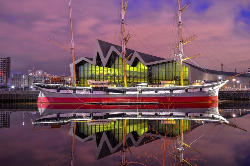 A large, red ship on the river with the pink sunset reflected in the water.