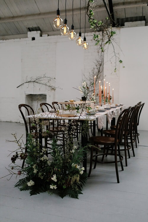  A long wedding table with greenery and candles in a sparse, white room.