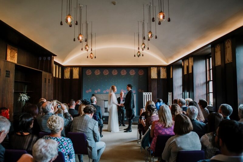 a couple hold hands getting married at the front of an art deco room