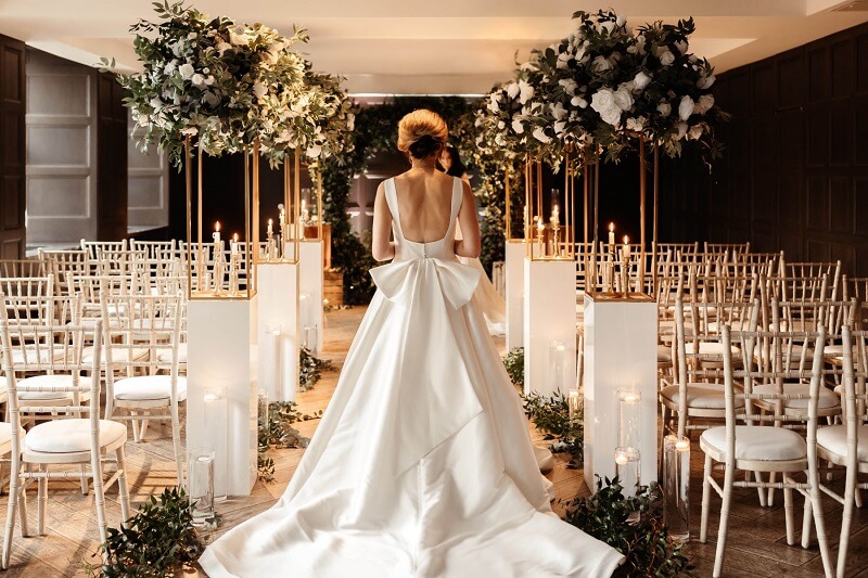 Bride wearing a wedding gown in the middle of an empty ceremony room decorated with white flowers.