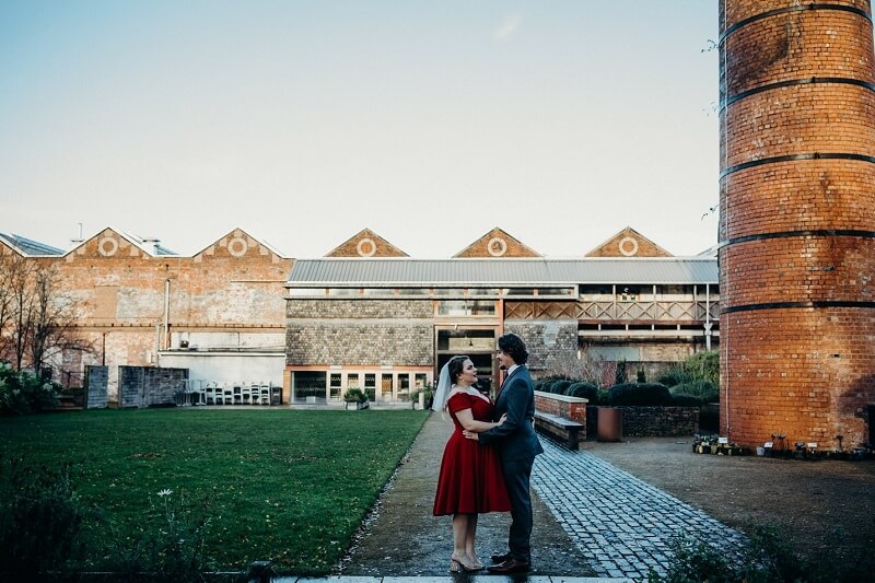 Bride and Groom holding each other in front of the wedding venue. Bride wears a bright Red dress and white wedding vale. Groom is wearing a blue suit.