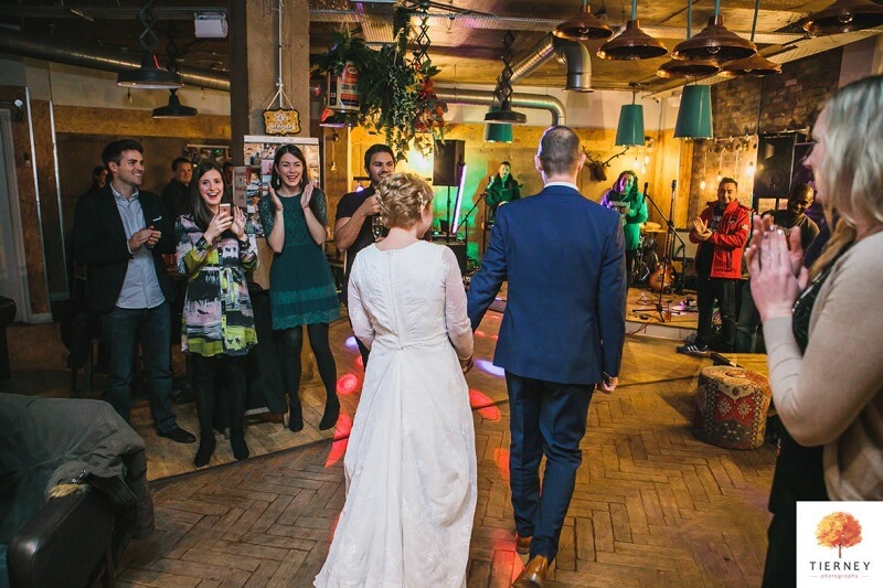 Bride and Groom walking in the middle of their reception room with wedding guests all around cheering