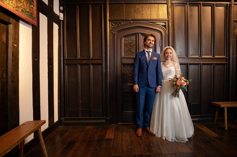 Cermeony Room with Wooden Floor and Wall. Bride and Groom standing holding hands and smiling. Bride is holding a bouquet of pale pink roses in her left hand. 
