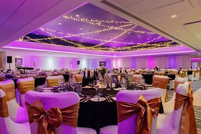 Wedding Hall at the OEC dressed for a wedding with black table cloth, white chair covers with gold sashes and a purple light ambience