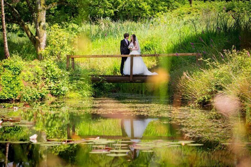 Bride and Groom standing on a wooden bridge going across a water pond. Plenty of green trees and foliage in the background 