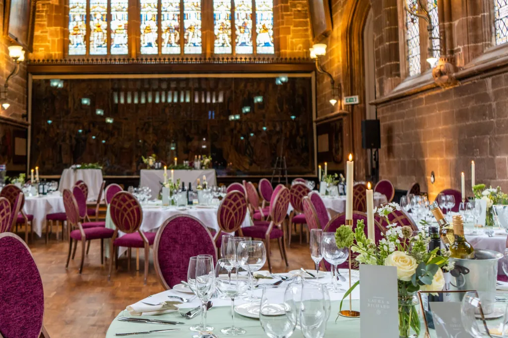 Wedding reception Room layes out with roundtables with white cloths and lit candles. Burgundy chairs around the table. 