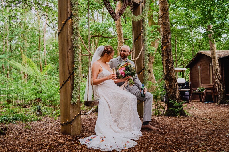 Bride and Groom sitting on a swing seat. the Bride is wearting a white dress and holding a bouquet of flowers in her left hand and a glass of champagne in the right hand. Groom is in a silver suit and holding a bottle of beer in his hand. Wooden Forest Background with a wooden cabin.