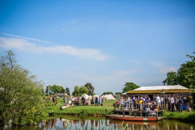 Outdoor wedding Reception by a little lac with blue sky day. Wedding Guests can be seen enjoying themselves around a marquee. 